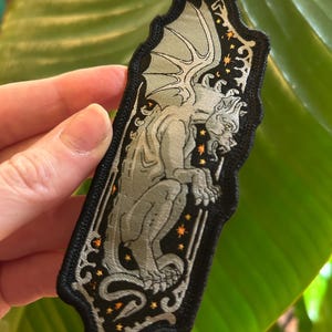 May include: Embroidered patch featuring a grey gargoyle with outstretched wings, set against a black background with orange star accents. The patch has a decorative border and is rectangular with rounded edges.