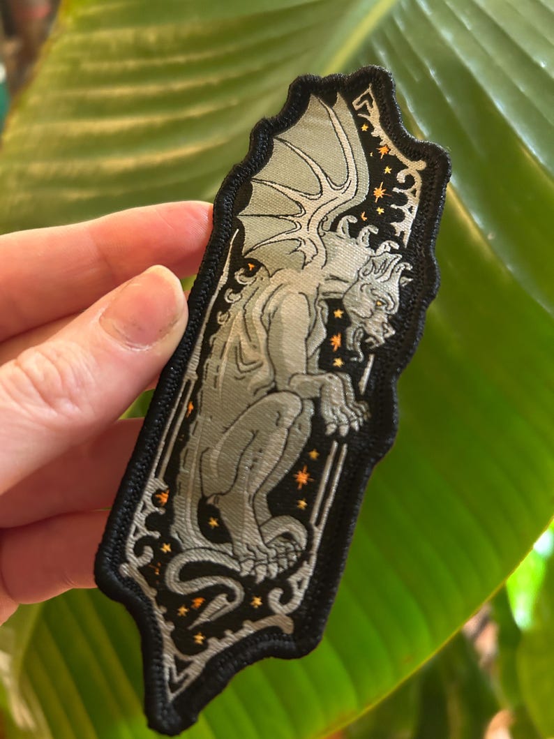 May include: Embroidered patch featuring a grey gargoyle with outstretched wings, set against a black background with orange star accents. The patch has a decorative border and is rectangular with rounded edges.