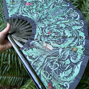 May include: A folding hand fan featuring a black base with an intricate light green botanical pattern of swirling leaves and vines. The design includes detailed illustrations of various beetles and insects scattered throughout the foliage.