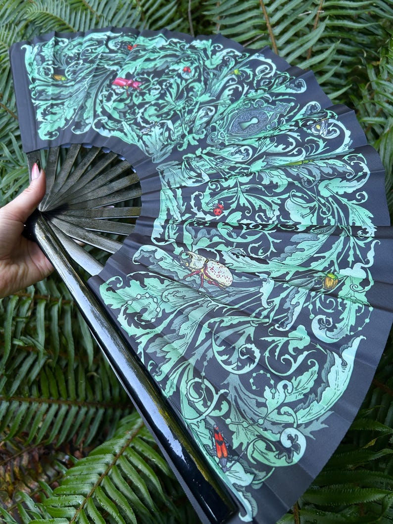 May include: A folding hand fan featuring a black base with an intricate light green botanical pattern of swirling leaves and vines. The design includes detailed illustrations of various beetles and insects scattered throughout the foliage.