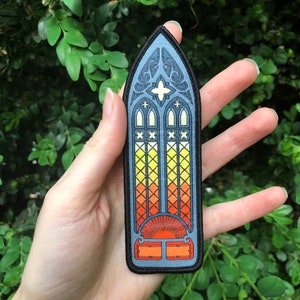 May include: A blue and black embroidered patch featuring a stained glass window design. The window is divided into three sections with a yellow, orange, and red gradient. The window has a black frame and a cross at the top.