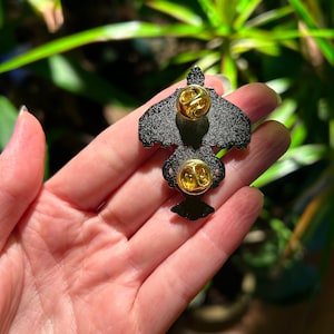 May include: Black enamel pin with a gold-colored butterfly design. The pin has two gold-colored butterfly clasps on the back.