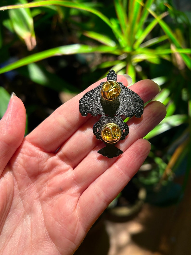 May include: Black enamel pin with a gold-colored butterfly design. The pin has two gold-colored butterfly clasps on the back.