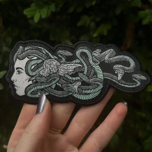 Puede incluir: Un parche bordado en blanco y negro que representa a Medusa con serpientes como cabello. Las serpientes son verdes y blancas y la cara está detallada con un contorno blanco.