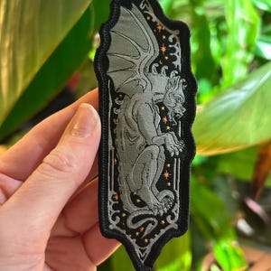 May include: A grey gargoyle patch with a black border. The gargoyle is detailed with wings and a fierce expression. The background is black with small orange star-like accents. The patch is rectangular with a decorative top and bottom.