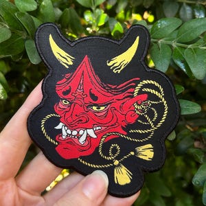 May include: A red and black embroidered patch featuring a Japanese demon mask with yellow horns and a gold rope with tassels.