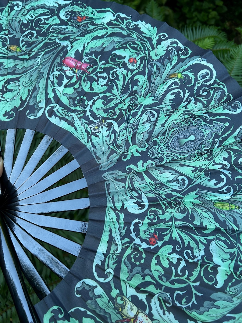 May include: A folding hand fan with black ribs and a dark fabric leaf featuring a light green botanical pattern of swirling leaves, various beetles, and a central keyhole motif. The design includes small red ladybugs and detailed insect illustrations.