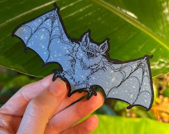 Puede incluir: Un parche azul bordado para planchar con una silueta de murciélago sobre un fondo de cielo estrellado. El murciélago está volando con las alas extendidas y tiene un diseño de telaraña.