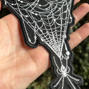 May include: Black and white embroidered patch of a spider web with a spider hanging from it.