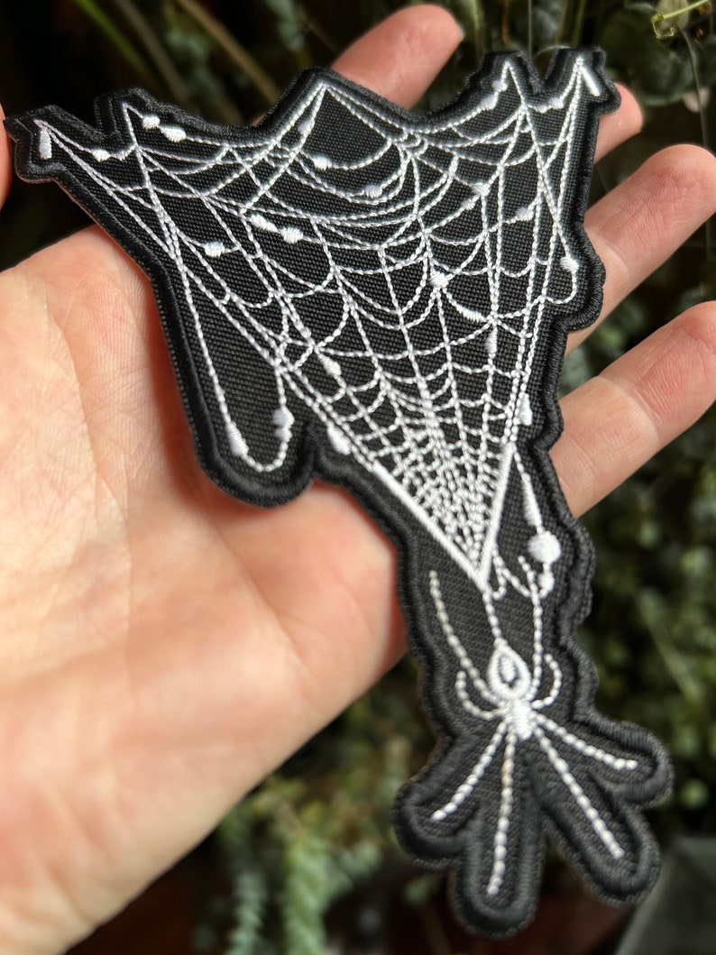 May include: Black and white embroidered patch of a spider web with a spider hanging from it.