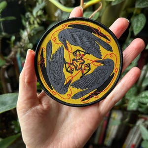 May include: Round patch with a yellow background featuring three black crows arranged in a triangle. Red lines connect the crows and a central symbol. The patch has a black border with red stitching, suitable for jackets, bags, or other fabric items.