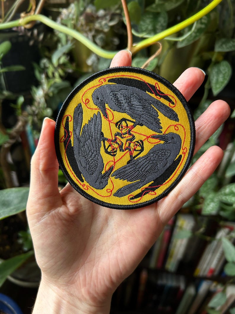 May include: Round patch with a yellow background featuring three black crows arranged in a triangle. Red lines connect the crows and a central symbol. The patch has a black border with red stitching, suitable for jackets, bags, or other fabric items.