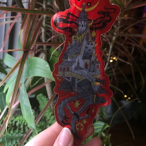 Puede incluir: Una pegatina roja con una ilustración detallada de un edificio gótico, una luna creciente y estrellas. El edificio es gris con detalles dorados, sobre un fondo rojo con elementos negros en forma de humo.