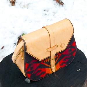 Pode incluir: Uma bolsa de couro marrom com fecho de aba e um padrão tecido colorido na frente. A bolsa tem uma fivela de metal dourado e uma alça de metal dourado.