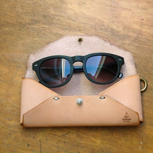 May include: A tan leather sunglasses case with a flap closure and a metal clasp. The case has a triangle logo with the text "BLOSS" printed on it. A pair of black sunglasses with dark lenses is inside the case.
