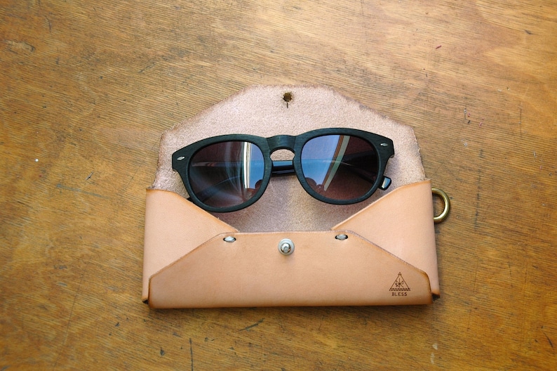 May include: A tan leather sunglasses case with a flap closure and a metal clasp. The case has a triangle logo with the text "BLOSS" printed on it. A pair of black sunglasses with dark lenses is inside the case.