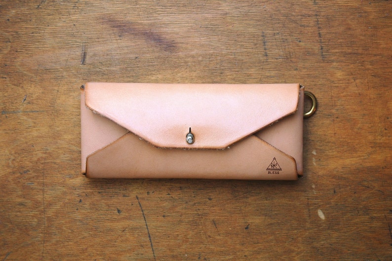 May include: A light brown leather wallet with a flap closure and a small metal snap. The wallet has a triangle logo with the text "Aomori Bless" on the front.