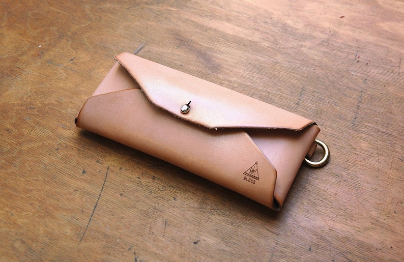 May include: A light brown leather wallet with a flap closure and a metal snap. The wallet has a triangle logo with the word "Bless" printed inside.