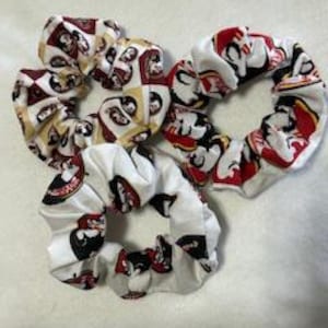 May include: Set of three white scrunchies with a red, yellow, and black patterned design. The pattern features a mascot with a helmet and a number.