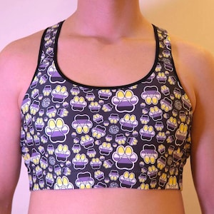 May include: A racerback sports bra featuring a repeating pattern of paw prints on a black background. Each paw print is divided into three horizontal sections colored yellow, white, and purple, outlined in white with black trim along the neckline.