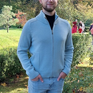 Pode incluir: Um homem vestindo um suéter azul claro com zíper e jeans azul está em um parque com folhagem de outono ao fundo.