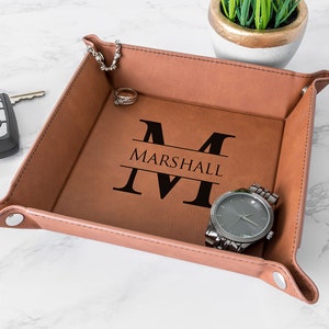 May include: A brown leather valet tray with the name "Marshall" and initial "M" engraved in black. A watch, keys, rings, and coins are inside the tray. A small plant in a gold and white pot is in the background.