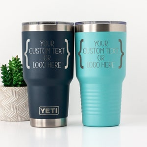 May include: Two stainless steel tumblers with a navy blue and a turquoise blue finish. Both tumblers have a silver lid and a silver base. The tumblers are engraved with the text "Your custom text or logo here" in a silver font. The tumbler on the left has the word "YETI" engraved on the base.