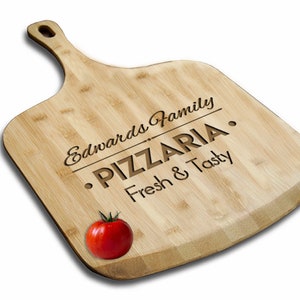 May include: A wooden pizza peel with a handle, engraved with the text "Edwards Family - PIZZERIA - Fresh & Tasty". A red tomato is sitting on the peel.