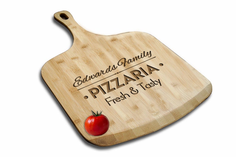 May include: A wooden pizza peel with a handle, engraved with the text "Edwards Family - PIZZERIA - Fresh & Tasty". A red tomato is sitting on the peel.