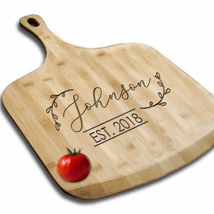 May include: A natural bamboo cutting board with a handle, engraved with the text "The Johnson EST. 2018" in a cursive font with a leafy design. A red tomato is in the foreground.