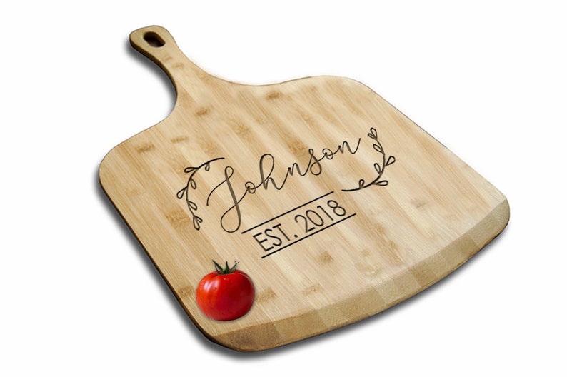 May include: A natural bamboo cutting board with a handle, engraved with the text "The Johnson EST. 2018" in a cursive font with a leafy design. A red tomato is in the foreground.