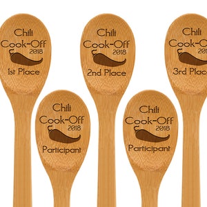 May include: Five wooden spoons with engraved text and a chili pepper graphic. The spoons are labeled "Chili Cook-Off 2018" with placements: 1st, 2nd, 3rd, and two "Participant" spoons. The spoons are made of light-colored wood.