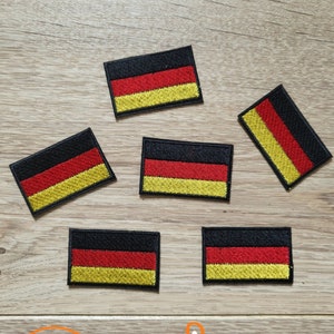 May include: Six black, red, and yellow embroidered German flag patches on a wooden surface. The patches are rectangular and have a black background with a red stripe and a yellow stripe. The text "Anja Creation Broderie" is visible at the bottom of the image.