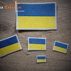 May include: Five embroidered patches of the Ukrainian flag. The patches are different sizes and are arranged in a row on a wooden surface. The largest patch is at the top of the image and the smallest patch is at the bottom. The patches are all white with a blue top half and a yellow bottom half.