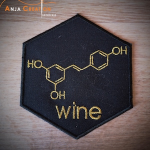 Puede incluir: Un parche hexagonal negro con bordado dorado de una fórmula química para el vino. El texto "Wine" también está bordado en oro.