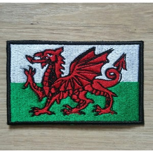 May include: Embroidered patch featuring the Welsh flag. The design includes a red dragon on a white and green background, with a black border. The patch is rectangular and likely for application on clothing or accessories.