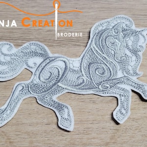 Peut inclure: Un patch brodé argenté représentant une licorne avec des détails complexes. La licorne est tournée vers la gauche et a une longue crinière et une longue queue flottantes. Le patch est sur une surface en bois brun clair.
