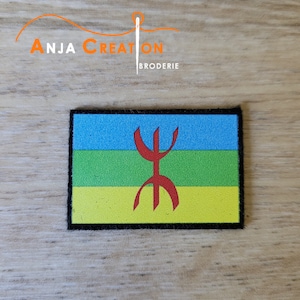 Peut inclure: Un patch brodé bleu, vert et jaune avec un symbole rouge au centre. Le patch a une bordure noire et est sur une surface en bois.