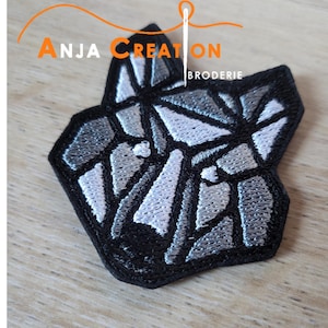 Peut inclure: Un patch brodé géométrique noir et blanc représentant la tête d'un loup. Le patch a un contour blanc et est brodé avec du fil gris et blanc. Le texte "Anja Creation Broderie" est visible en arrière-plan.