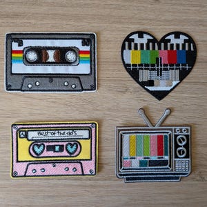 May include: Four embroidered patches featuring retro media themes: a grey cassette tape, a heart-shaped television test pattern, a pink and yellow cassette tape labeled "Best of the 90's", and a grey television set with an antenna and color bars.