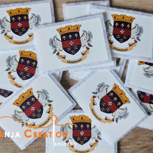Peut inclure: Gros plan sur plusieurs patchs en tissu blanc avec un blason coloré représentant un écu, une couronne et deux oiseaux. Le blason est entouré d'une bannière avec le texte "QUANALAO".