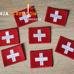 May include: Eight red embroidered iron-on patches with a white Swiss cross design. The patches are on a wooden surface.