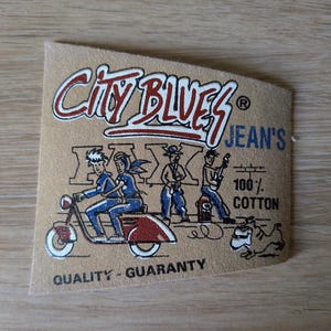 Könnte beinhalten: Ein beiges, rechteckiges Kleidungsetikett mit dem Text City Blues Jean's und Quality-Guaranty. Die Grafik zeigt zwei Personen auf einem roten Roller, zwei stehende Figuren und einen Hund. Das Etikett enthält zudem den Text 100% Cotton in Schwarz.