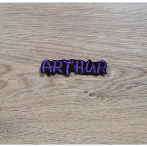 Puede incluir: Parche bordado morado con el nombre "ARTHUR" en una fuente estilizada.