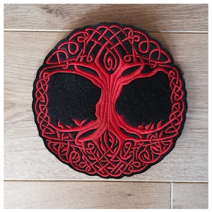 Puede incluir: Diseño de árbol de la vida celta bordado en rojo sobre un fondo de fieltro negro. El árbol está rodeado por un borde de nudos.