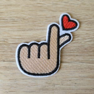 May include: Embroidered patch featuring a hand gesture forming a heart shape. The patch has a tan hand with black outlines and a red heart on a white background. The patch is designed to be ironed on or sewn onto fabric.