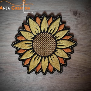 Könnte beinhalten: Ein gestickter Sonnenblumen-Patch in Gelb und Orange mit brauner Mitte. Der Patch befindet sich auf einem Holzgrund.