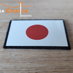 Op de afbeelding: Een witte patch met een rode cirkel in het midden, die de vlag van Japan voorstelt. De patch is omgeven door een zwarte rand en is gemaakt van stof.