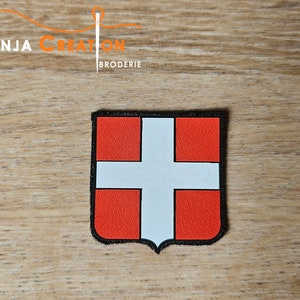 May include: A red and white embroidered patch with a white cross on a red background. The patch is shaped like a shield and has a black border.