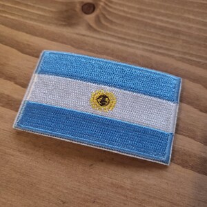 Può includere: Toppa ricamata raffigurante la bandiera argentina. Il disegno presenta tre strisce orizzontali: due strisce azzurre in alto e in basso e una striscia bianca al centro. La striscia centrale presenta un emblema del sole giallo.